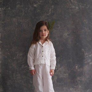 Pu&ograve; includere: Una bambina con una camicia e pantaloni di lino bianco sta in piedi davanti a uno sfondo grigio testurizzato. Ha i capelli lunghi e castani e indossa sandali marroni.