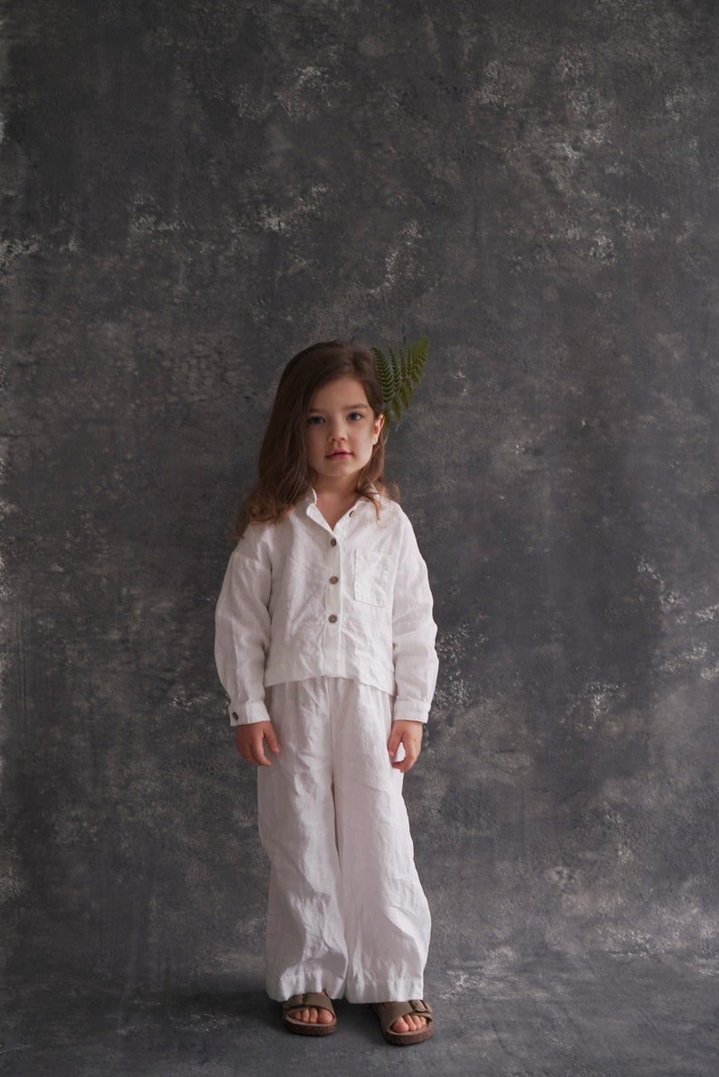 Pu&ograve; includere: Una bambina con una camicia e pantaloni di lino bianco sta in piedi davanti a uno sfondo grigio testurizzato. Ha i capelli lunghi e castani e indossa sandali marroni.
