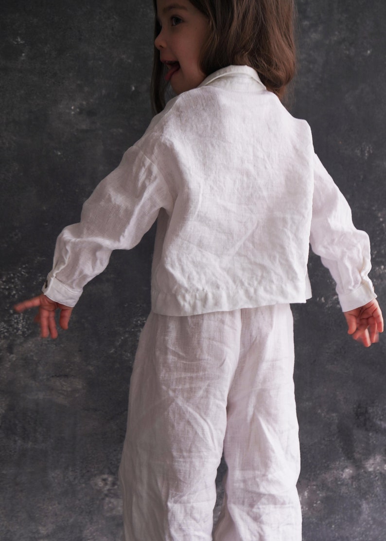 Pu&ograve; includere: Un completo di camicia e pantaloni in lino bianco per un bambino. La camicia ha un colletto classico e maniche lunghe. I pantaloni hanno una vestibilit&agrave; rilassata.