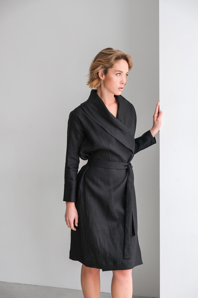 May include: A black linen wrap dress with a long sleeve and a tie belt. The dress has a wide collar and a relaxed fit.