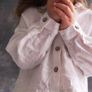 Pu&ograve; includere: Un bambino piccolo che indossa una camicia bianca di lino con bottoni marroni. Il bambino tiene una foglia di felce verde davanti al viso.