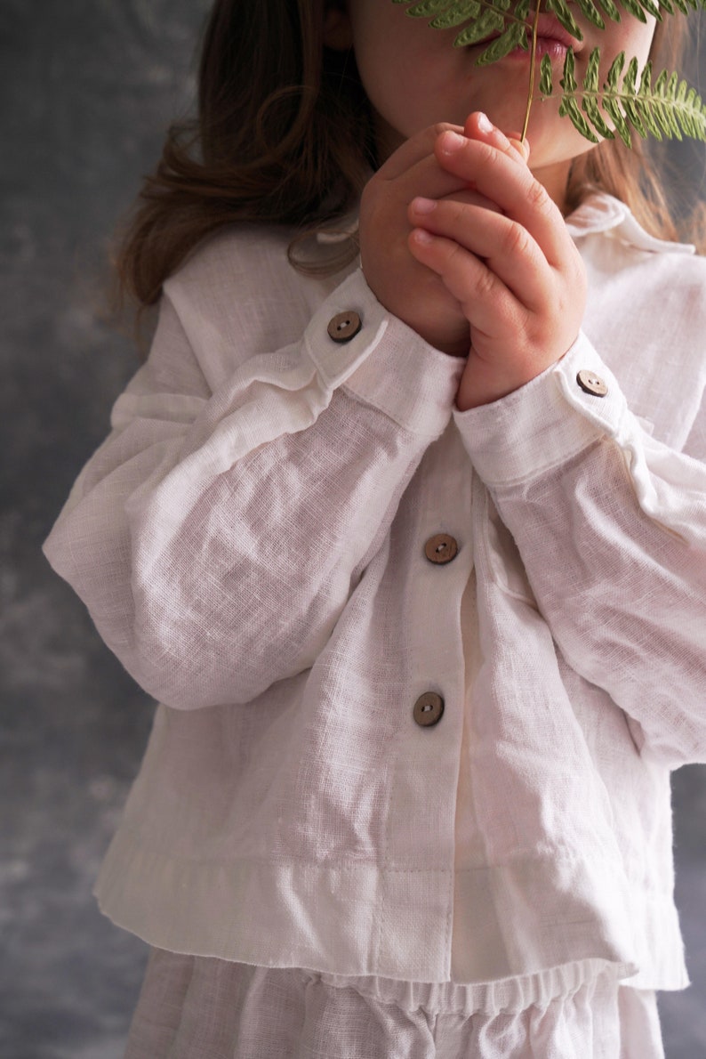 Pu&ograve; includere: Un bambino piccolo che indossa una camicia bianca di lino con bottoni marroni. Il bambino tiene una foglia di felce verde davanti al viso.
