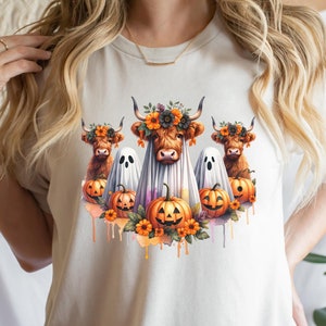 May include: A white t-shirt with a graphic of three Highland cows dressed as ghosts for Halloween. The cows are wearing flower crowns and surrounded by pumpkins and fall foliage.