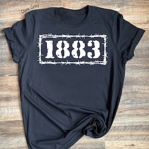 May include: A dark gray t-shirt with a white graphic of the number 1883 inside a barbed wire frame.