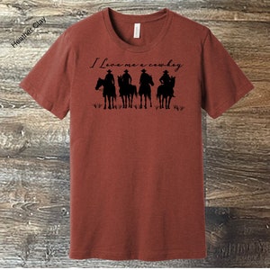 May include: A rust-coloured t-shirt with the text "I Love me a cowboy" printed in black. The design features four silhouettes of cowboys riding horses.