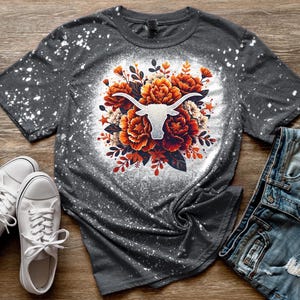 May include: A gray t-shirt with a white bleach-splattered design. The design features a white silhouette of a longhorn bull's head surrounded by orange and white flowers.