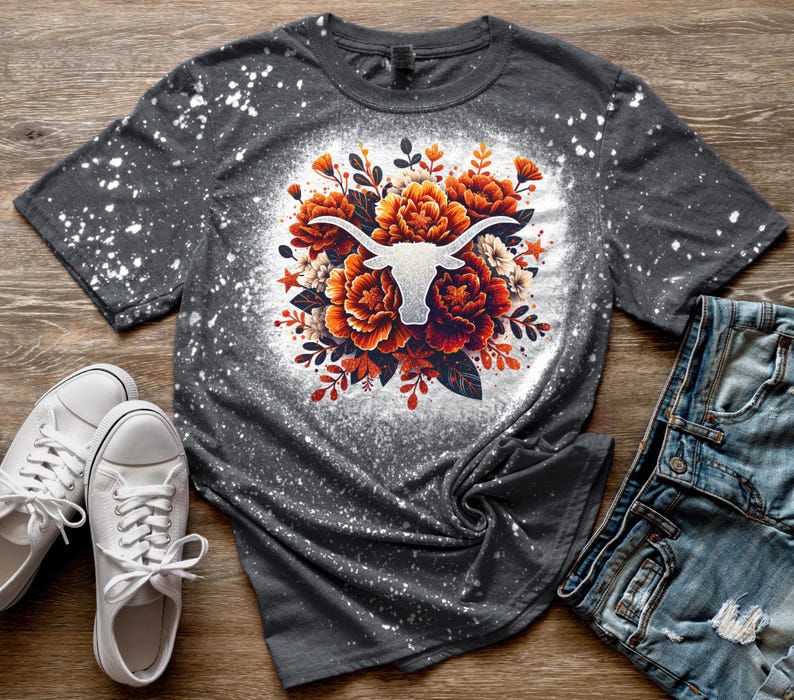 May include: A gray t-shirt with a white bleach-splattered design. The design features a white silhouette of a longhorn bull's head surrounded by orange and white flowers.