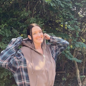 May include: A zip-up hoodie featuring a solid brown center panel and sleeves with a dark green, blue, and white plaid pattern. The garment includes a hood with matching brown drawstrings, a full-length front zipper, and two front pockets.
