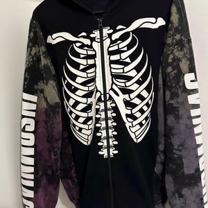 May include: A black zip-up hoodie featuring a white skeleton rib cage graphic on the front. The sleeves have a tie-dye pattern in shades of purple, grey, and black, with the word INSOMNIAC printed in white capital letters along the lower arms.