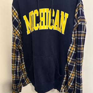May include: A navy blue hooded sweatshirt featuring the word MICHIGAN in large yellow block letters across the chest. The garment includes a front kangaroo pocket, a drawstring hood, and long sleeves made of a navy, yellow, and white plaid fabric.