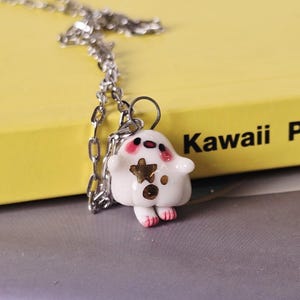Puede incluir: Un collar de cadena plateada con un colgante blanco con forma de personaje de dibujos animados con manchas marrones y pies rosas. El colgante cuelga frente a un libro amarillo con las palabras "Kawaii Party".