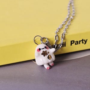 Puede incluir: Un collar con un colgante de gato de cerámica blanca y una cadena plateada. El gato tiene detalles en negro y dorado y patas rosas. El collar descansa sobre un libro amarillo con la palabra "Party" y un libro azul con la letra "S" y el nombre "Louisa Taylor".