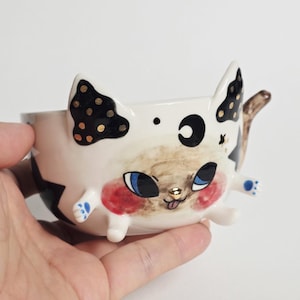 May include: A ceramic bowl shaped like a cat with pointed ears, featuring a cream-coloured base, black and gold polka-dot ears, a black crescent moon symbol, blue eyes, pink cheeks, a small gold nose, and tiny protruding paws on the sides.