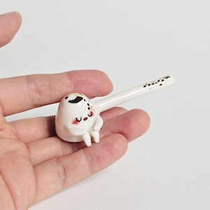 May include: A small white ceramic spoon rest featuring a stylized character with a rounded body, a smiling face with pink cheeks, and small legs. The handle is long and slender with a black and gold speckled pattern near the end.