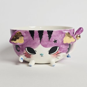 May include: A ceramic bowl shaped like a cat with a purple painted exterior, black stripes, and gold leaf accents on the ears. The bowl features a small cat tail handle, four small feet, and a painted face with large black eyes and pink cheeks.