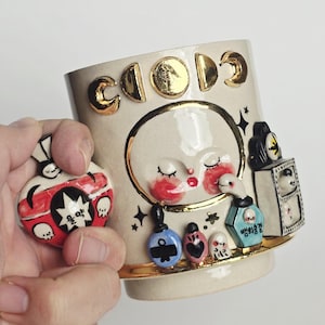 May include: Cream-colored ceramic mug with gold accents and a whimsical design. The mug features a face with closed eyes and red cheeks, along with various decorative elements. A small, red and white charm is held next to the mug.