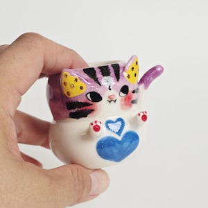 May include: A small ceramic cup shaped like a cat with a white base, purple and black striped head, and yellow spotted ears. The cat features a blue heart on its chest, small raised paws with red details, and a purple tail extending from the side.