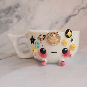 Könnte beinhalten: Eine weiße Keramiktasse mit einem skurrilen Weltraum-Design. Die Tasse hat ein Gesicht mit großen schwarzen Augen, rosa Akzenten und goldenen Verzierungen. Dekorative Sterne, Planeten und eine goldene Sonne zieren das Äußere und schaffen eine verspielte Ästhetik.