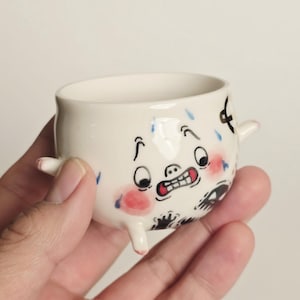 Op de afbeelding: Een kleine, witte keramische beker met een cartoongezicht. De beker heeft een wijd openstaande uitdrukking met rode wangen, een grijnzende mond en blauwe, traanachtige accenten. Hij heeft kleine, stompe benen en armen.