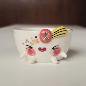 May include: A small white ceramic bowl featuring a hand-painted face with pink cheeks, a red mouth, and closed eyes. The bowl is decorated with gold star accents, a pink circular detail with a gold star, and a yellow striped element on the rim.
