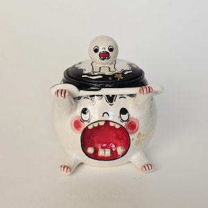 May include: A whimsical, handmade ceramic sugar bowl with a cartoonish monster design. The white bowl features a large open red mouth, pink cheeks, and small feet. The black lid is decorated with a white ghost-like figure and gold accents.
