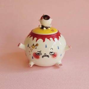 May include: A round ceramic jar with a lid featuring a small figure on top. The jar is white with a red drip pattern at the rim, blue teardrop accents, and a distressed facial expression. It has small arms and legs, with gold-colored details.