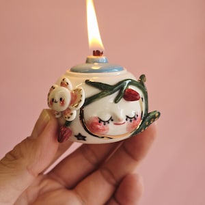 May include: A whimsical ceramic oil lamp with a lit wick. The lamp features a smiling face with closed eyes, rosy cheeks, and a crescent moon design. Adorned with floral and koala bear accents, the lamp is held against a pink background.
