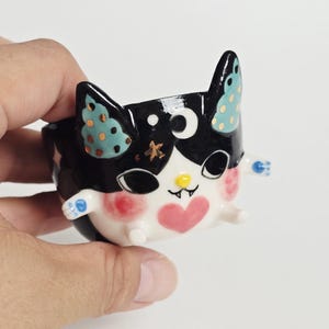 May include: A small ceramic cup shaped like a black and white cat with pointed ears, teal inner ears with gold dots, a white crescent moon on the forehead, a pink heart on the chin, rosy cheeks, a yellow nose, and tiny white arms and legs.