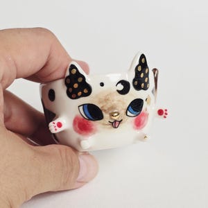 May include: A small white ceramic bowl shaped like a cat with black and gold spotted ears, blue eyes, pink cheeks, and a gold nose. The bowl features small protruding paws with red dots, a crescent moon symbol, and a tiny tongue sticking out.