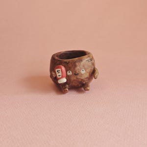 Puede incluir: Una maceta de cerámica marrón caprichosa con forma de personaje, que sostiene una pequeña botella de ketchup. La maceta tiene una cara sonriente y brazos y piernas pequeños. La botella de ketchup tiene el número "27".