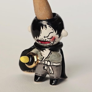 May include: A whimsical ceramic incense holder shaped like a stylized figure with black hair and a black cape. The figure holds a gold-colored ring and a black bag. An incense cone is placed in the head.
