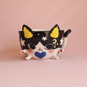 May include: A ceramic cat-shaped mug with a black and white design, featuring yellow ears with polka dots, a blue heart, and star and moon accents. The mug has a tail and small arms and legs, set against a pink background.