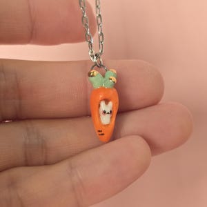 Könnte beinhalten: Halskette mit silberner Kette und Karottenanhänger. Der orangefarbene Karottenanhänger hat grüne Blätter und einen kleinen weißen Hasen im Inneren. Die Halskette wird vor einem rosa Hintergrund präsentiert.