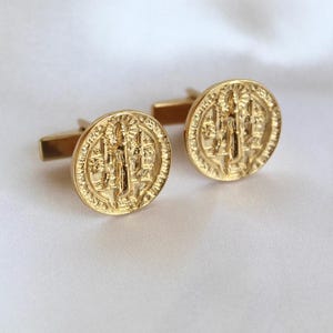May include: A pair of gold-colored cufflinks with a detailed circular design. Each cufflink features a raised relief image of a religious figure and text. The cufflinks are set against a white background.