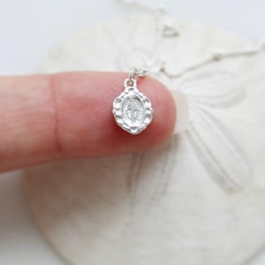 Peut inclure: Un petit collier pendentif en argent avec un charm ovale détaillé. Le charm a une surface texturée et un motif central. Le collier est présenté sur une surface blanche.