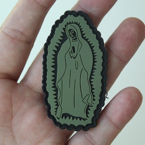 May include: An olive green patch featuring an illustration of the Virgin Mary. The patch has a black scalloped border and is held in a hand. The image of Mary is in a simple, elegant style.