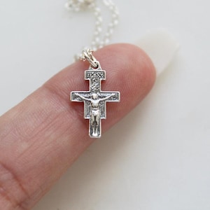 May include: A small, silver-colored crucifix pendant on a delicate chain. The cross features a detailed depiction of Jesus Christ. The pendant is held between two fingers against a white background. The jewelry is likely a religious item.