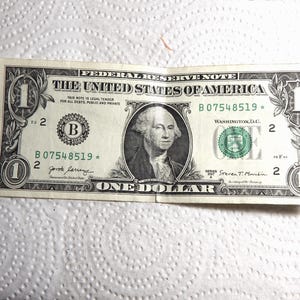 Puede incluir: Un billete de un dólar estadounidense, con un retrato de George Washington. El billete muestra el texto "THE UNITED STATES OF AMERICA" y "ONE DOLLAR". El número de serie es B07548519.