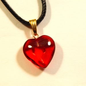 Può includere: Una collana con ciondolo a forma di cuore rosso. Il ciondolo è rosso traslucido e lucido, sospeso a un cordino nero. La cauzione è color oro. La collana è un accessorio romantico.