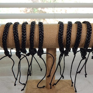May include: A display of black braided bracelets with adjustable ties. The bracelets are arranged on a tan, cylindrical display stand. The background includes a window with blinds.