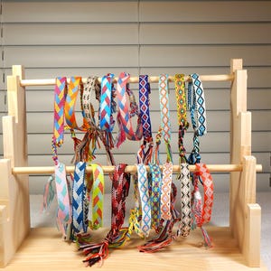May include: A wooden display stand holds a collection of colorful friendship bracelets. The bracelets feature various patterns and color combinations, including blue, red, yellow, and green. The stand is made of light-colored wood.