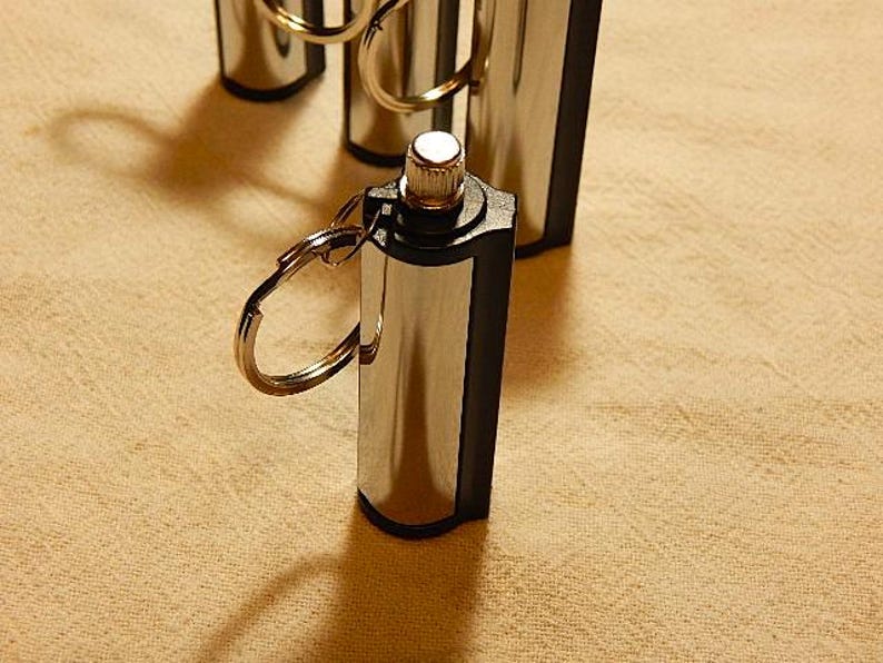 May include: A silver and black metal fire starter with a key ring. The fire starter has a small, round, silver button on the top.