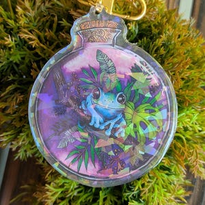 May include: A clear, round keychain featuring a blue frog surrounded by green and purple foliage inside a glass jar. The keychain has a gold-colored clasp and is set against a backdrop of green moss.