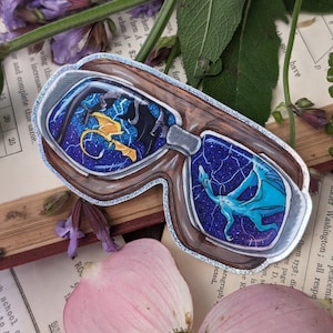 May include: A sticker featuring a pair of brown-framed glasses with a fantasy scene inside each lens. One lens shows a yellow dragon, the other a blue dragon, both against a starry, glittery background. The sticker has a silver glitter border.