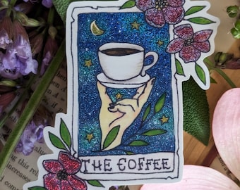 May include: A sticker with a glittery blue background featuring a hand holding a cup of coffee. The sticker has a white border and the text "THE COFFEE" at the bottom. The sticker is decorated with pink flowers and green leaves.