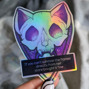 May include: A holographic sticker of a cat skull with a purple and blue gradient. The sticker has a quote that reads "If you can't summon the flames directly from hell store bought is fine."