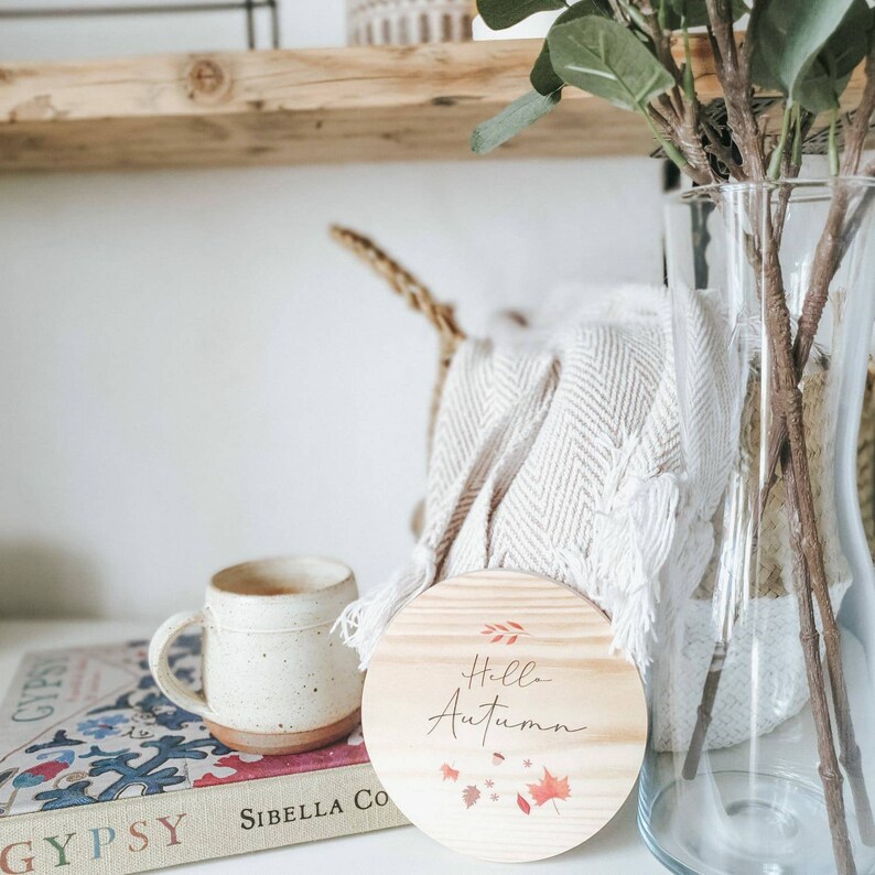 May include: A ceramic mug with a speckled finish and a wooden coaster with the text "Hello Autumn" and leaf illustrations. The mug is next to a woven basket and a glass vase with branches.