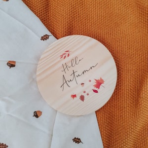 May include: A circular wooden sign with the text "Hello Autumn" in black script, adorned with red and orange leaves and acorns. The sign rests on a white cloth patterned with acorns and leaves, against an orange textured backdrop.