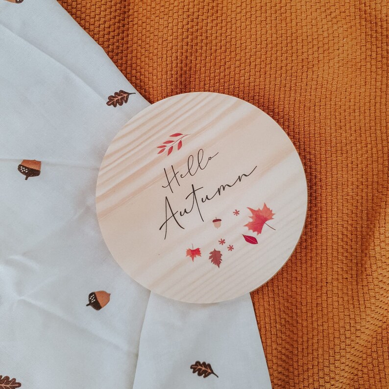 May include: A circular wooden sign with the text "Hello Autumn" in black script, adorned with red and orange leaves and acorns. The sign rests on a white cloth patterned with acorns and leaves, against an orange textured backdrop.