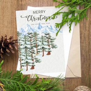 May include: A Christmas card with a watercolor illustration of snow-covered pine trees and mountains. The card reads "MERRY Christmas AND HAPPY NEW YEAR" in elegant script. The card is inside a kraft paper envelope, with pine needles and a pine cone on a wooden surface.